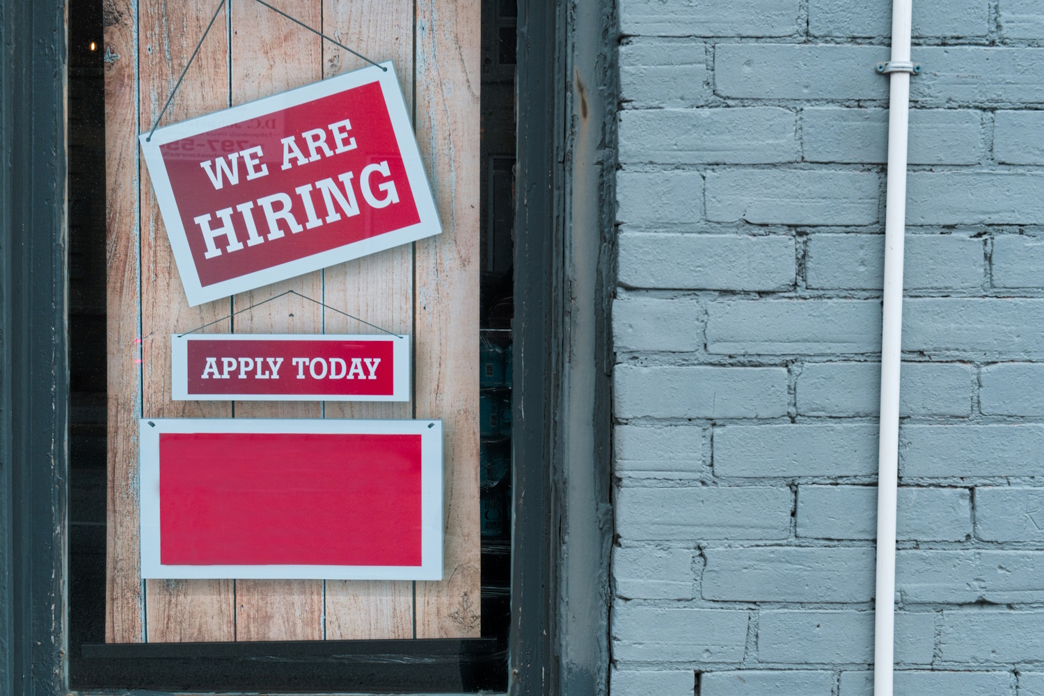ID: two rectangular signs in a boarded up display window next to a brick wall, both written in white capitalised letters on a red background. The first, hanging askew, reads: "We are hiring". The second, below, reads: "Apply today".